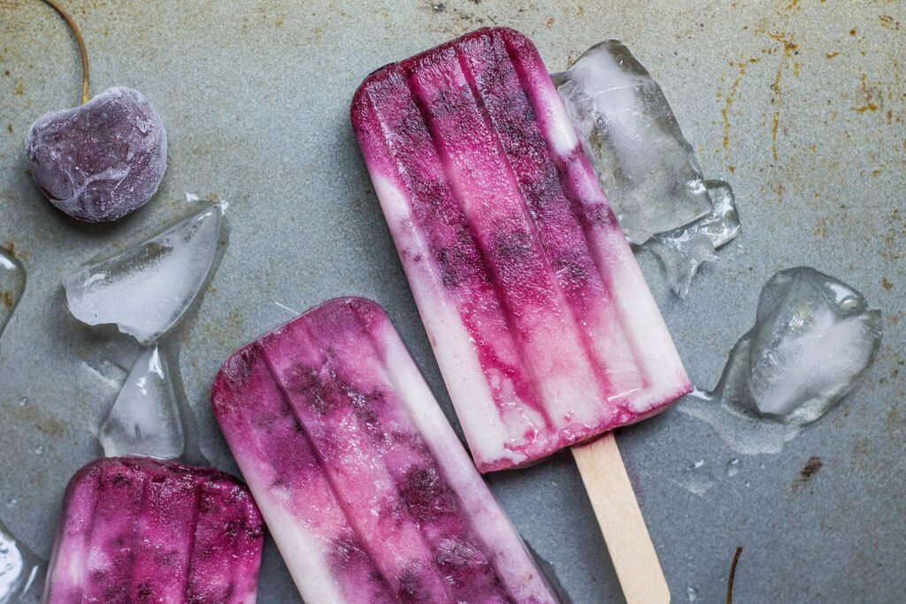 Three Queen Garnet yoghurt swirl popsicles sitting on a table, with glass and frozen berries alongside