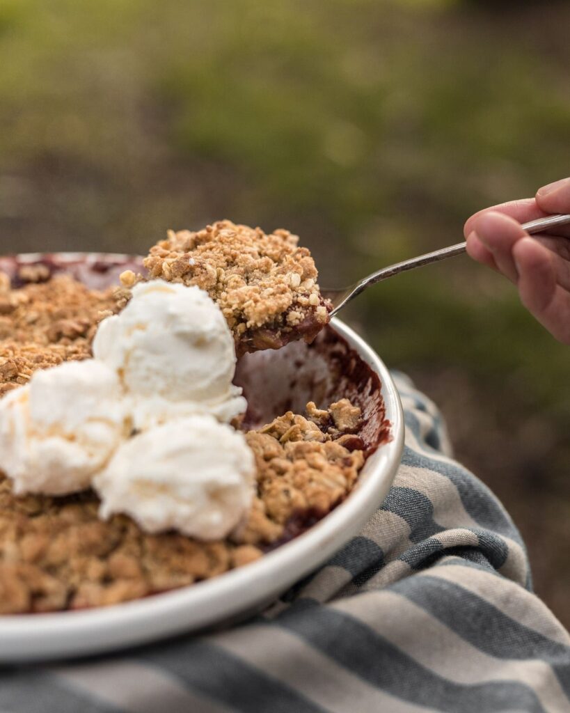 Plum + Apple Crumble topped with cream being spooned out to serve